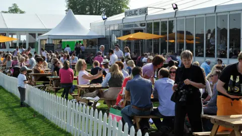 Badminton Horse Trials Crowds sit and drink and chat in front of a white picket fence at an open air bar with a white marquee in the sunshine