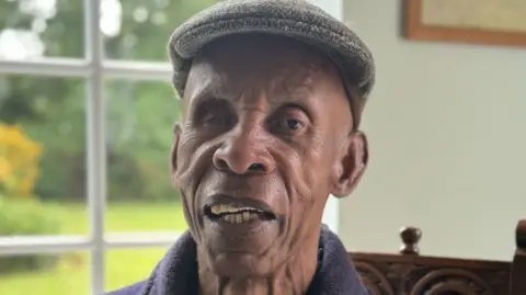A man wearing a flat cap and blue fleece is sitting inside his house by a window. He is sitting on a wooden chair, with the garden of grass and flower beds visible through the white-framed window.