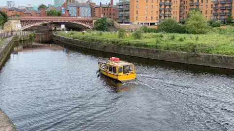 A yellow motor boat is on a river sailing towards a bridge, surrounded by tall buildings 