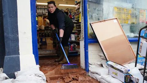 Woman sweeps brown muddy water out of a shop door with a gap made in sandbags over the front step