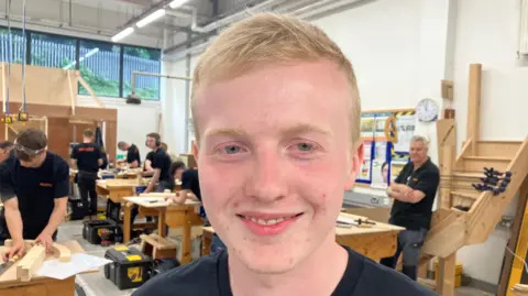 David Magee has brown hair and blue eyes and is wearing a black t-shirt. He is standing in front of other people in black t-shirts doing wood-work.