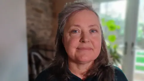 Paula Richards in her early 50s, with long hair, looking at the camera, in her home