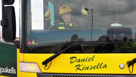 Gaskells Daniel as a child sits in the driver's seat of a yellow bin lorry named after him. He is wearing a hard hat and holding two thumbs up while smiling.