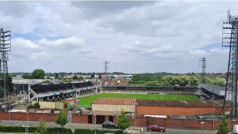 LDRS A city football stadium, with the pitch in the middle surrounded by stands. In each corner of the stadium is a tall flood-light structure.