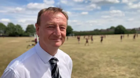 Simon Spark/BBC A man standing in front of children playing football. He has short brown hair and is wearing a green and grey striped tie and a white shirt.
