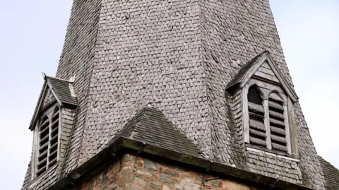 A close up shot of the lower part of a church spire, back by a blue sky. There are small oak panels covering the spire, with wooden, worn through windows.