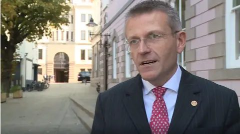 Philip Ozouf stood on the right wearing a suit. He has a pinstriped blazer with a red and white tie. He is looking to the left and is talking. In the background are buildings, one with an arched doorway.