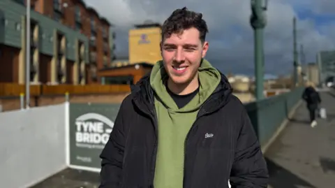 A young man wearing a green hoodie and a black coat standing on the pavement which goes across the Tyne Bridge 