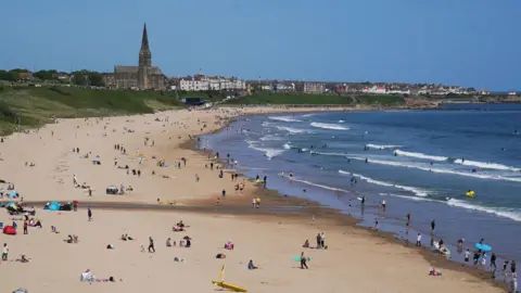 PA Media Tynemouth's Longsands beach on a sunny day. It has a number of people on the sand and in the water - but not hoardes. The waves are making white breakers near the shore and the skyline of Cullercoats is in the background.