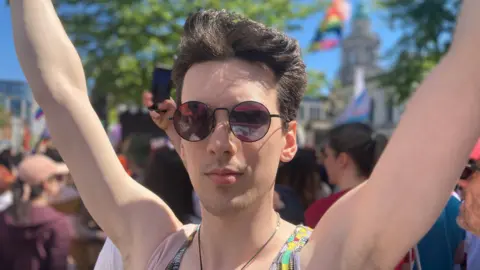 Autumn has short brown hair and round dark sunglasses on. Behind him is the city hall and blurred protesters.
