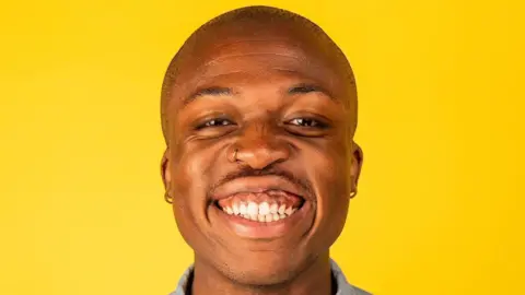 Abena Kusi Benjy is bald and is smiling at the camera. He has a gold nose ring and hoop earrings. He is standing in front of a bright yellow background. 