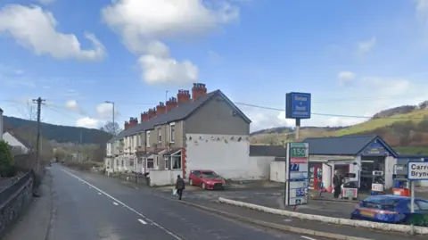 A5 near Llidiart-y-Parc in Denbighshire. A garage can be seen off the road on the right hand side. Behind the garage there are a row of terraced houses. A person can be seen walking along the road. A blue sky is in the background. 