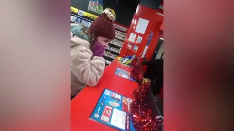 Eileen Mitchell A teenage girl is covering her ears with her fingers. She wearing gloves, a hat and a cost. She's standing at the cash register in a shop. Tinsel is decorating the counter top. Shelves of DVDs are in the background.