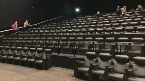 Shariqua Ahmed/BBC Inside one of the cinema rooms. There are many rows of padded black seats, gradually getting higher up towards the back of the room. A few people are browsing in the rows. The walls are black.