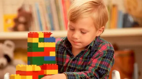 The image shows a young child seated at a table, building a tall structure out of colourful interlocking plastic bricks.