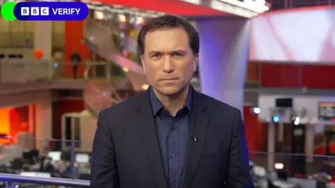 A man, Ros Atkins, wearing a dark blue suit stands with a newsroom behind him