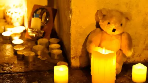 PA Candles, a teddy bear, and a holy water font lie on the ground.
