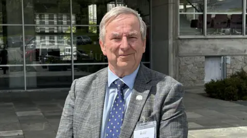A man with grey hair in a grey jacket with a blue shirt and dark blue tie standing in front of County Hall in Truro