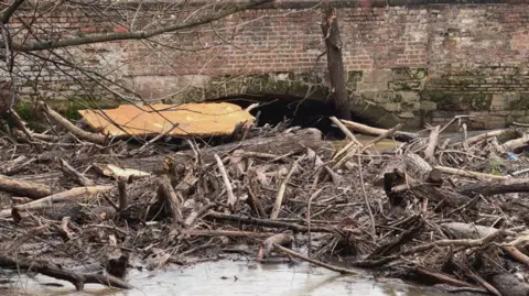 Piles of tree branches in a large river, butting up against a bridge structure. 