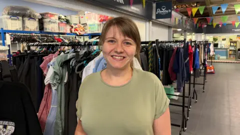Ashleigh Hirst standing in the charity's uniform store, smiling at the camera. She has brown hair tied back and a fringe. She is wearing a green T-shirt.