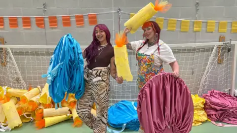 Two women standing in a hall holding up homemade bright yellow flaming candles and blue and pink costumes. The woman on the left is wearing animal print trousers and a brown top and has long dark pink-tinted hair. The woman on the right has red dyed long hair in pigtails and is wearing a white T-shirt and bright floral dungarees. Piles of candles and costumes are behind the pair, in a mouth of a football goal.