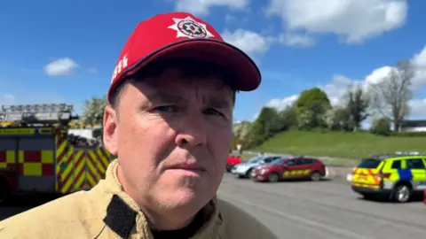 David Doherty has short dark hair and is wearing a red cap with the Northern Ireland Fire and Rescue badge on it. He is wearing a brown firefighter suit. Behind him is a yellow and red fire engine and a yellow and blue police car along with other vehicles.