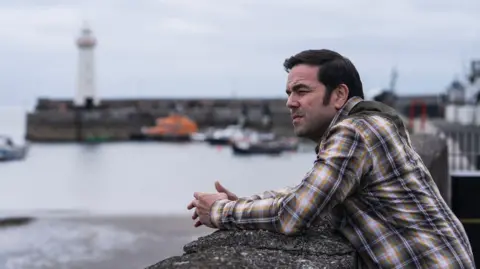 BBC/Long Story TV/Peter Marley A man is pictured leaning on a sea wall. He's dressed in a brown and white check shirt and has dark hair. The background is blurred but you can see boats and the lighthouse. 