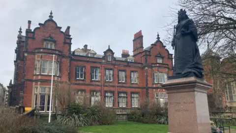 An outdoor scene featuring a large historic building made of red brick, along with a prominent statue in the foreground. The statue stands on a tall, polished stone pedestal. The statue depicts Queen Victoria wearing an elaborate crown-like headpiece and holding a sceptre. The pedestal includes engraved text: "VICTORIA REGINA 1837–1901" Behind the statue is a large red‑brick building with elaborate architectural details. Notable features include: Tall chimneys with decorative tops.
Multiple gables with ornate stonework.
Arched windows framed with darker stone.
A mix of rectangular and bay windows.
A slightly weathered appearance, consistent with an older historic structure. There is vegetation around the building, including bushes and small plants. A flagpole stands to the left of the building.