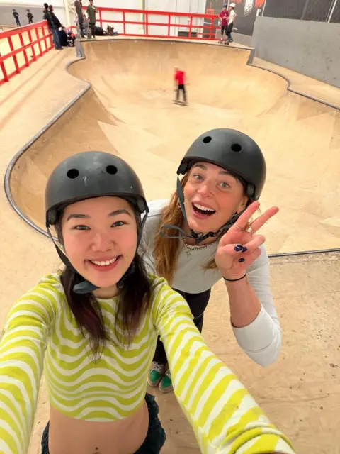 Winona Two girls in black helmets smile at the camera with a wooden skate park behind them