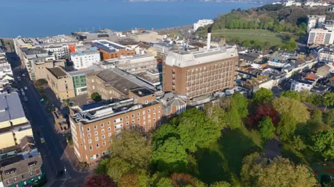 BBC A drone shot of Jersey General Hospital in St Helier. It's a large building amongst lots of other buildings and a green park with lots of trees. The sea is also visible in the background. 