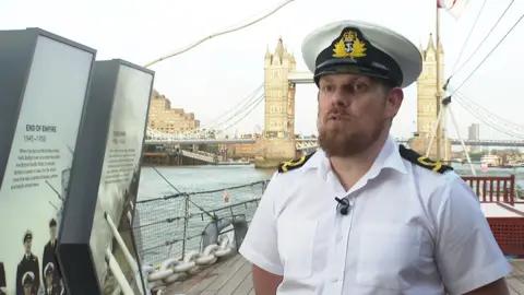 A man in a seaman's uniform aboard a ship in front of Tower Bridge