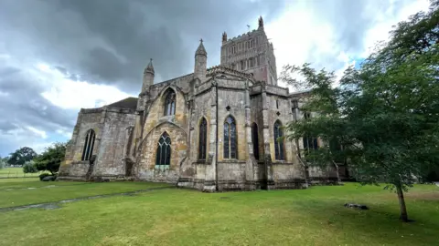 Tewkesbury Abbey, a large church, stands on a very green, cut lawn which has a couple of trees on it. There is a grey, cloudy sky above it with a few patches of sunlight illuminating some clouds.
