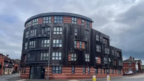 A four storey residential building, with a mix of brown brick, wooden cladding and windows sits on a roundabout next to two roads.