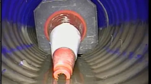 An orange and white traffic cone on its side inside a drain. It is lying in a brown liquid. 