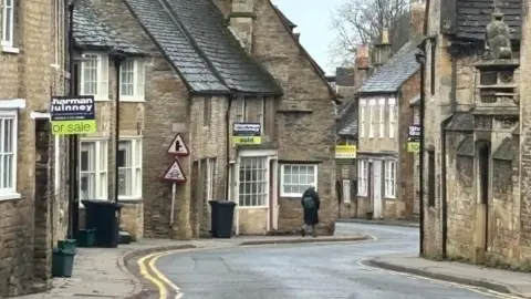 Brian Farmer/BBC North Street, Oundle: A grey haired woman wearing a black coat and backpack walks down a winding street which has light brown stone houses on either side.