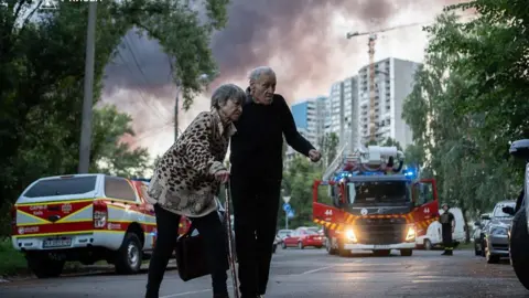 An elderly woman and man with ambulances and smoke in the sky behind them