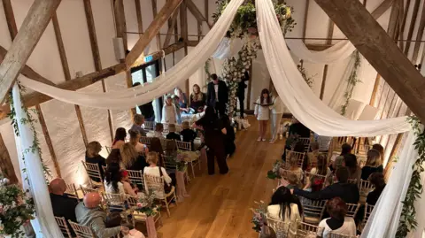 A wooden hall with flowers and many people. It is a wedding. 
