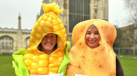 Katie Harris is standing on the left and is wearing a costume which is a piece of sweetcorn. Ruth Nye is on the right and is wearing a potato costume. They are both standing next to each other outside of an old building.