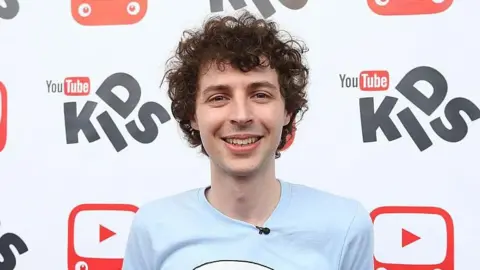 Getty Images YouTuber Stampy, who is smiling and looking at the camera. He has black curly hair and is young-looking, wearing a bright colourful pastel blue t-shirt. Behind him is a backdrop with the logo for YouTube Kids repeated.