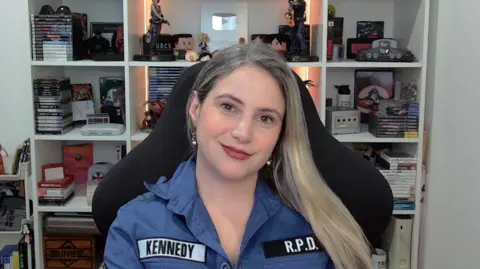 Monique Alves Monique Alves, a content creator who is smiling at the camera sitting in her computer chair, wearing a blue police shirt which says "Kennedy" and "RPD" on it. Behind her are shelves of Resident Evil memorabilia such as statuettes and games.