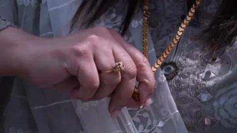 Maryam holding a thick yellow gold chain. There is a gold band on her finger with a silver stone.