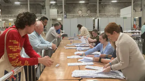 BBC People behind a table counting ballot papers, with several people looking on