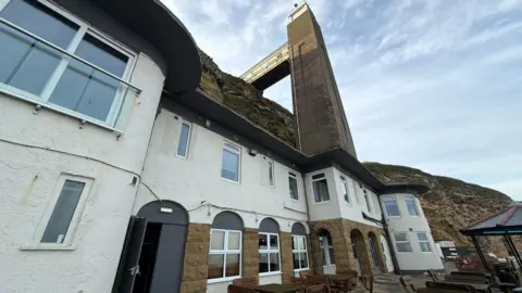 The Grotto is a two-storey venue with a white, black and brown stone exterior. A tall concrete lift shaft rises out of the building and up the cliff face. A walkway connects the top of the lift to the top of the cliffs.