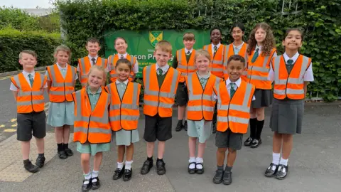 Fourteen pupils, with orange bibs over their uniforms, stand outside the school gates. The school sign can be seen behind them.