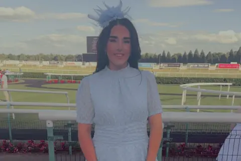 Supplied Ibbie wearing a light blue dress and a fascinator. She is stood outside with a race track behind her. 