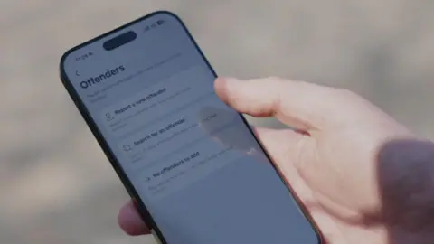 Close-up of a hand holding a smartphone displaying the new app, allowing users to "report a new offender" or "search for an existing offender".