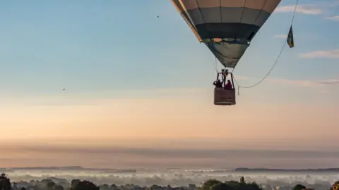 BBC Weather Watchers/Grum Two people inside a wicker hot-air balloon basket in the sky. One has a hand over their eyes like a shield. The sky in front of them is peach, blue and slightly grey with minimal cloud. Below them are treetops which are covered with white fog and mist