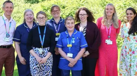 A group of eight people, some in health uniforms and some with lanyards. They are all smiling directly at the camera and they appear to be standing in a garden. There is a hedge behind them.