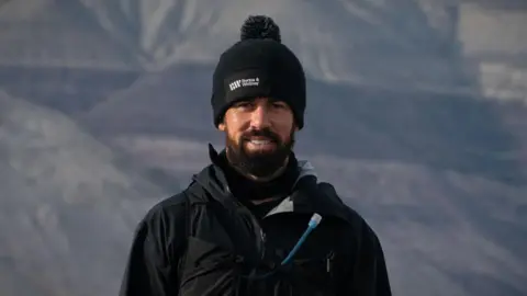 Juan Cruz Lopez A man with a dark brown beard and wearing a black woolly hat and jacket is standing looking directly into the camera. He is smiling with mountains in the background.