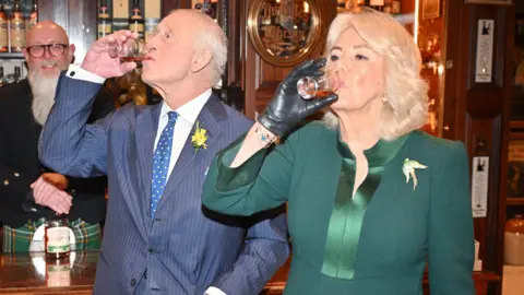 PA Media The King and Queen tasting whiskey in a wood panelled bar. The Queen is wearing a green dress with a brooch and she has blonde shoulder length hair. The King is wearing a blue pinstripe suit, a white shirt. He has white short hair. 